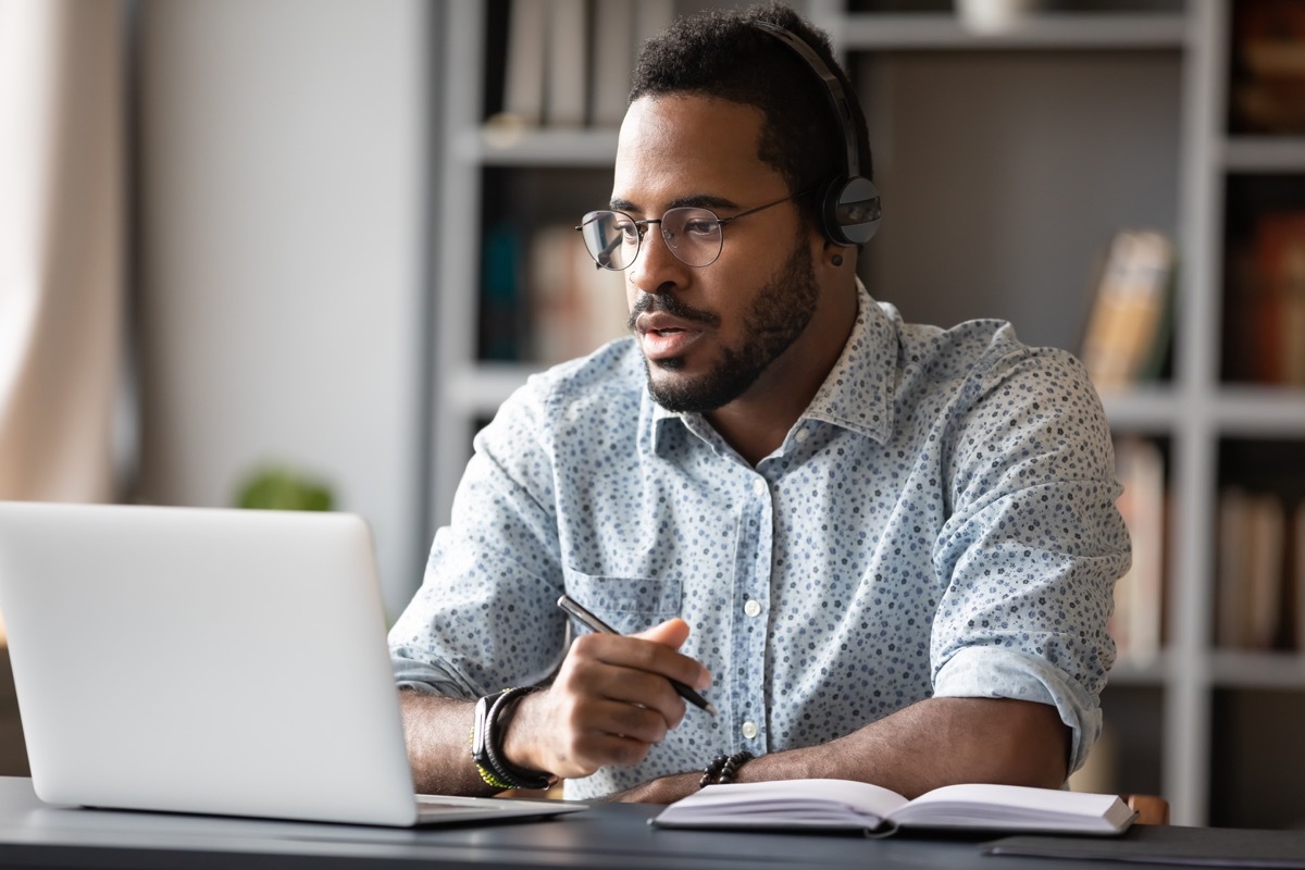 Focused young businessman wear headphones study online watching webinar podcast on laptop listening learning education course conference calling make notes sit at work desk, elearning concept