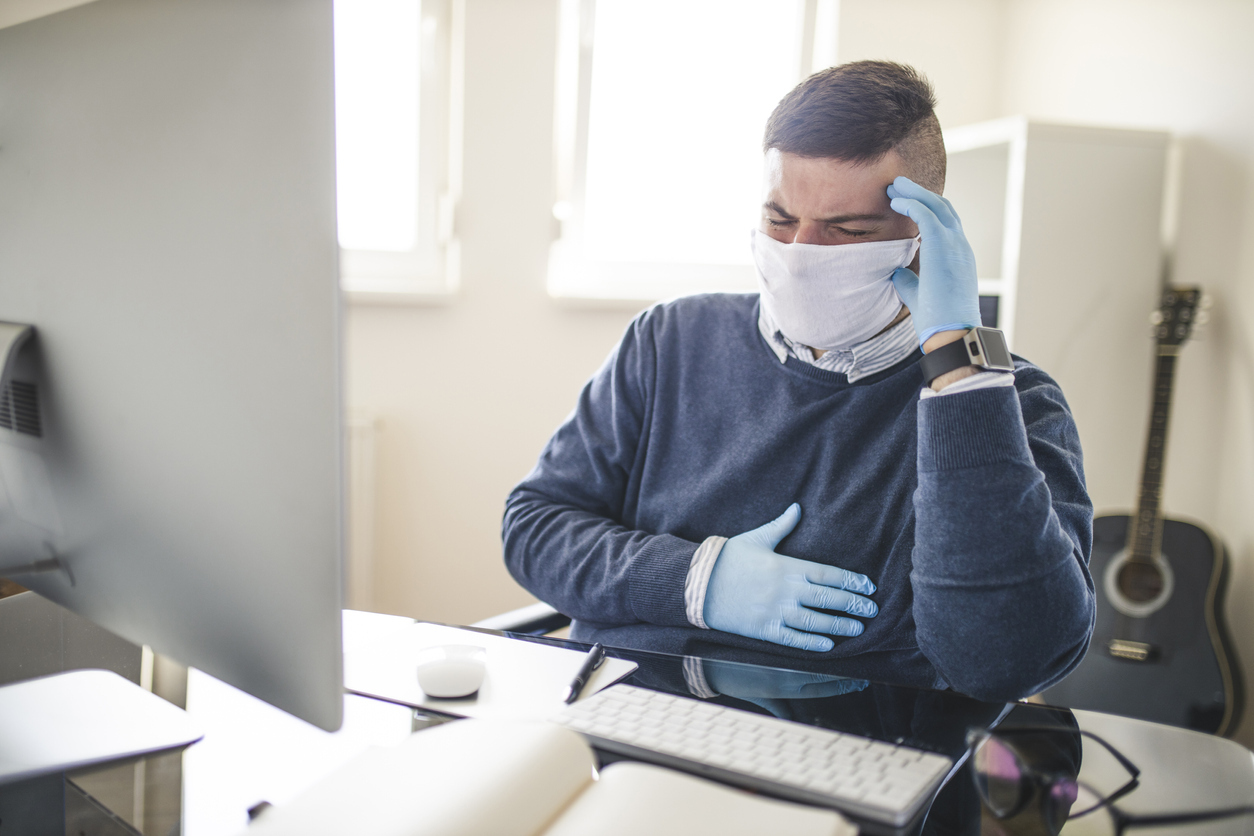 A young man sitting at a desk wearing a face mask and gloves grabbing his chest due