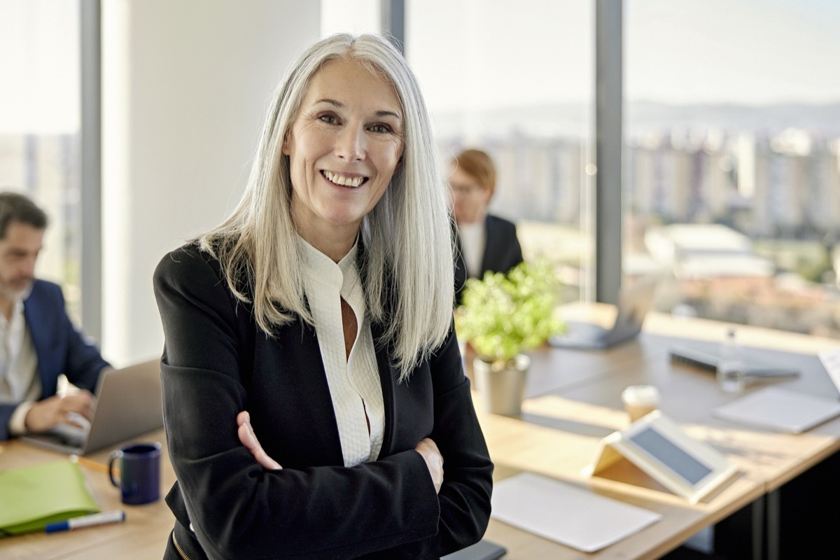 Waist-up front view of longhaired executive in businesswear sitting on table with arms crossed and smiling at camera as colleagues work in background.