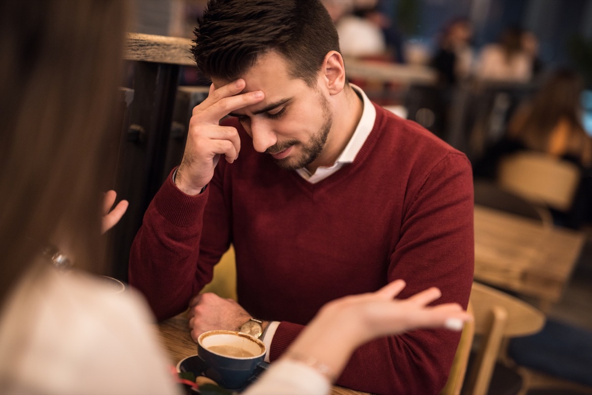 man looking down and touching his forehead while woman talks at a coffee shop