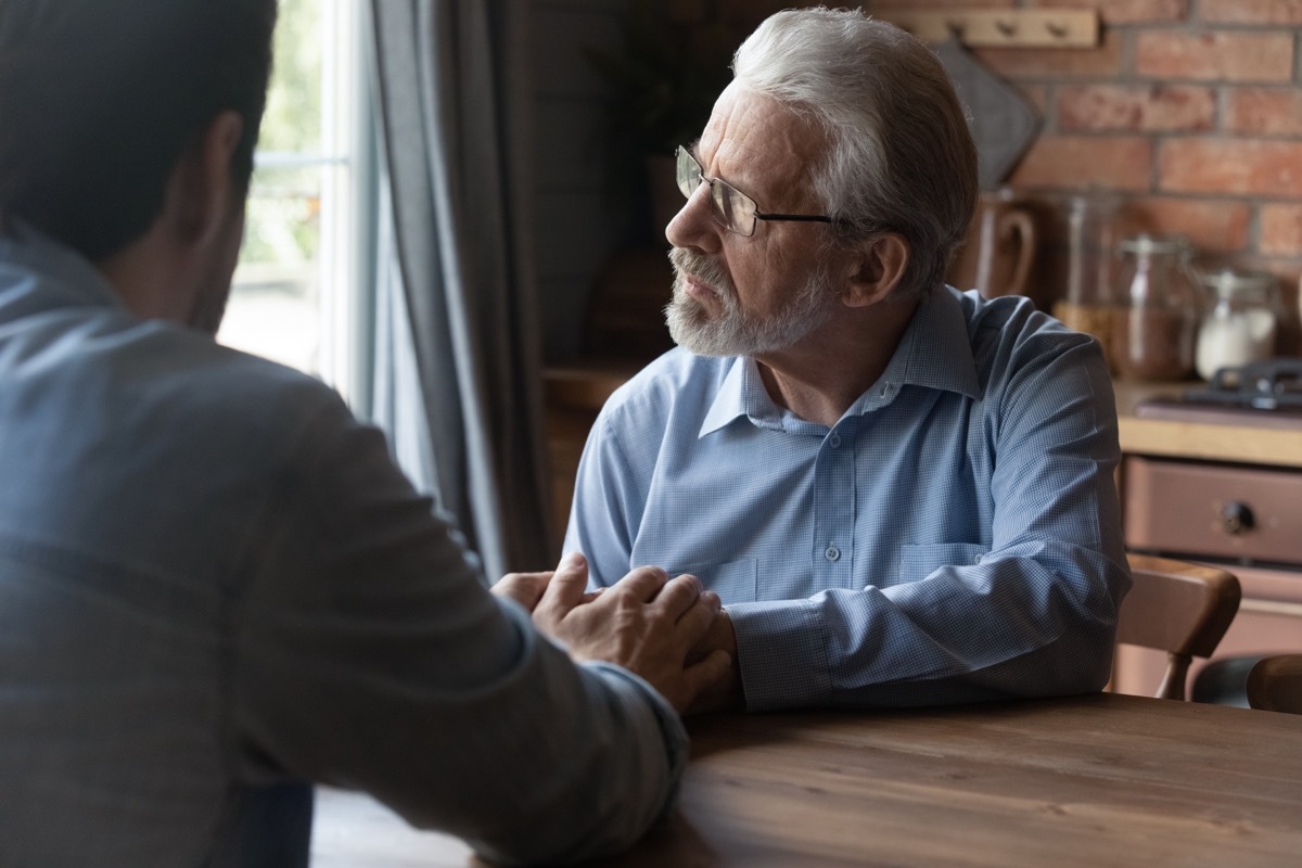 Helping hands of son. Attentive grownup kid sit at table close to sad senior hoary father touch his palms listen to problem. Young grandson give support to depressed old grandfather help overcome loss