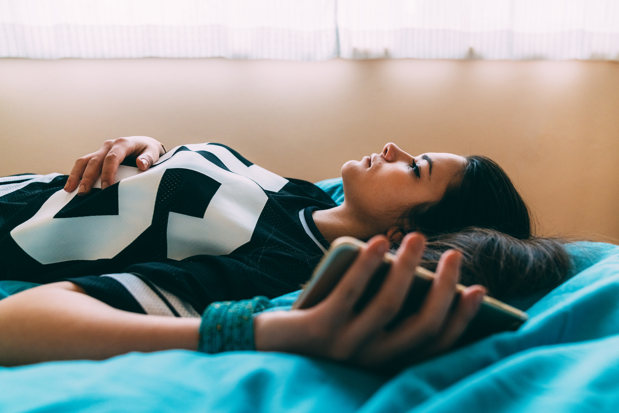 A young woman lies in bed while holding her phone with a depressed look on her face.