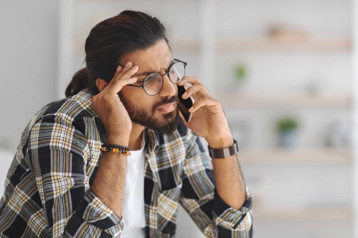 Upset middle-eastern guy in casual sitting on couch, having phone conversation with lover or business partner, experiencing difficulties