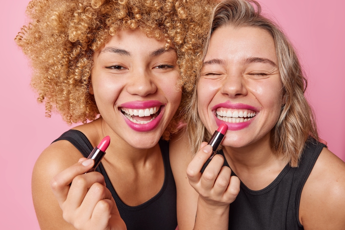 Close up of two friends smiling and putting on pink lipstick.