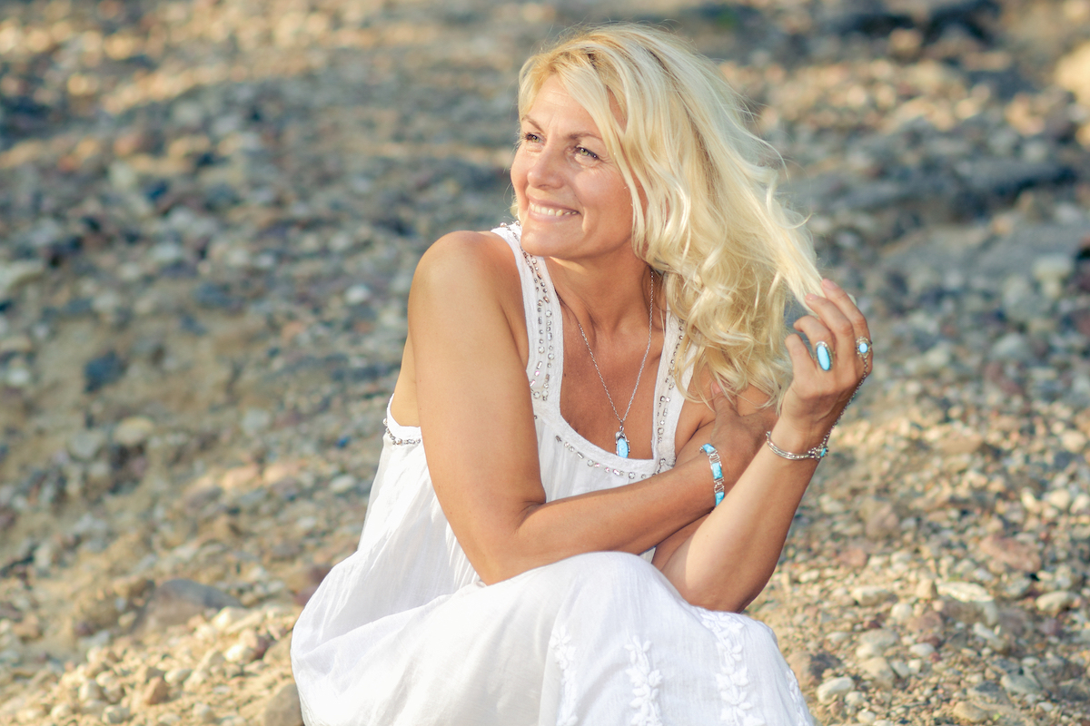 A middle-aged blonde woman wearing a white dress crouches down on a rocky part of the beach