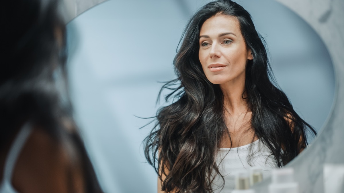 Beautiful Middle Aged Woman Looks into Bathroom Mirror, Enjoys Her Looks. Concept for Happiness, Wellbeing, Natural Beauty and Organic Skin Care Products