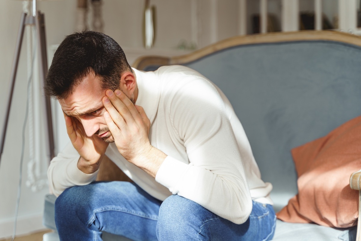 Sad male in a white pullover bending double with a pain on a sofa