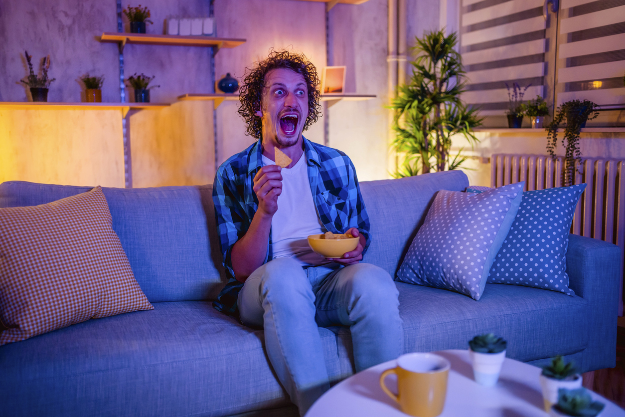 Young scared man watching horror movie and eating snacks in the living room at night