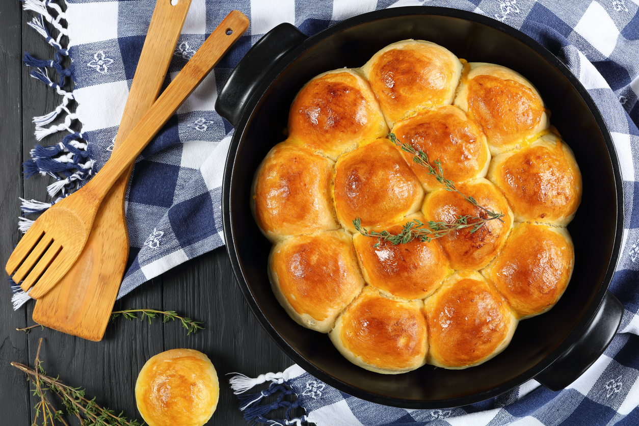 freshly baked soft and fluffy homemade dinner rolls in baking dish with kitchen towel and spatulas on wooden kitchen table, view from above, close-up