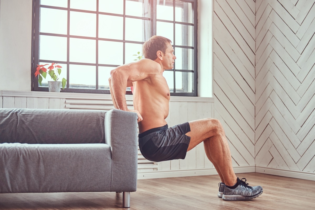 Handsome shirtless muscular male does exercise leaning on a sofa at home.