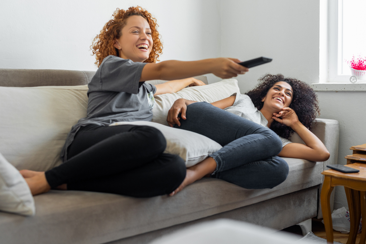 Two young women sitting on a couch using a remote and watching TV