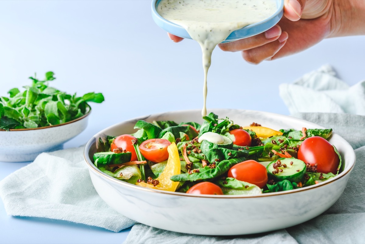 hand pouring a cream-based salad dressing onto a salad in a white bowl