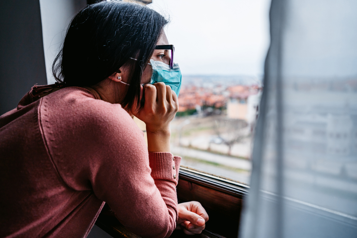 A young woman wearing glasses and a face mask looks out her window during a COVID lockdown