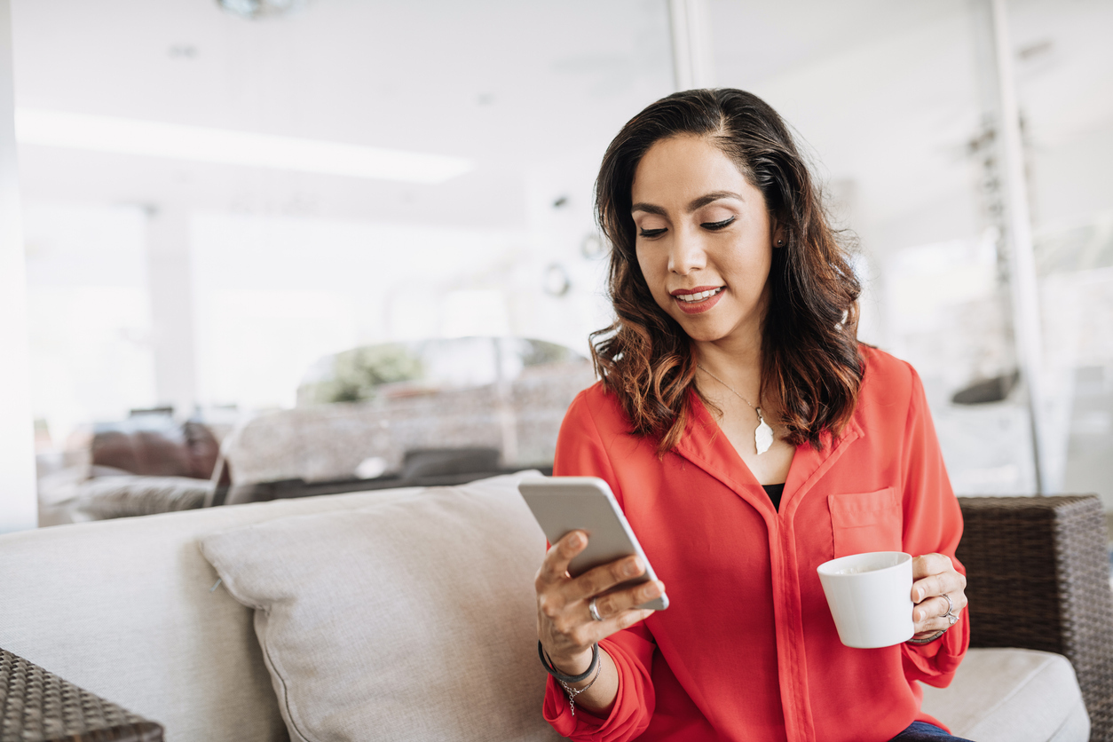 A young woman sitting on a couch checking her iPhone while holding a mug.