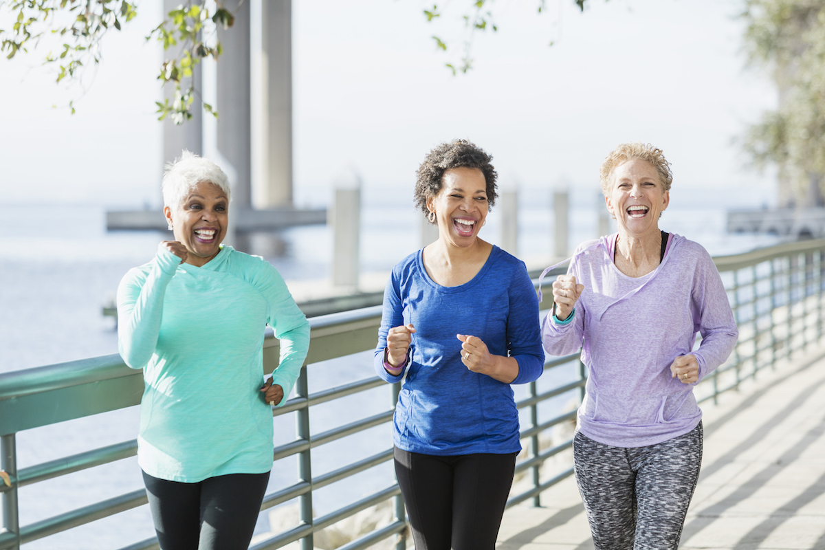 A group of three multi-ethnic women in their 50s and 60s power walking on a city waterfront side by side. They are having a good time, laughing.