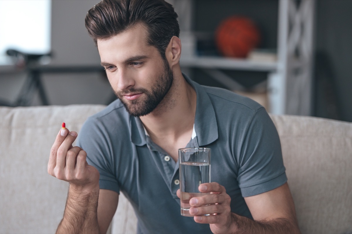 man holding a glass of water and looking at a pill in his hand while sitting on the couch