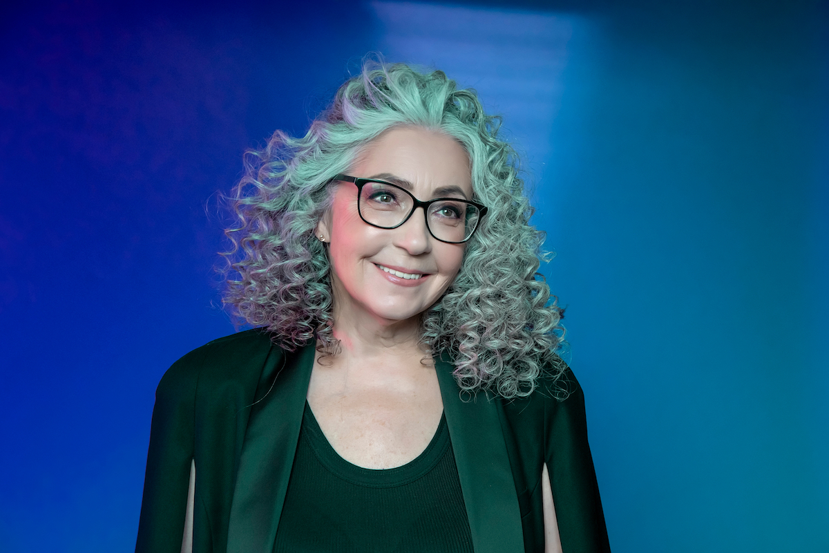 An older woman with curly gray hair wearing a green suit against a blue background.