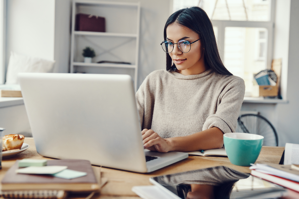 A young woman with long brown hair wearing blue glasses and a beige sweater is working on her laptop smiling.