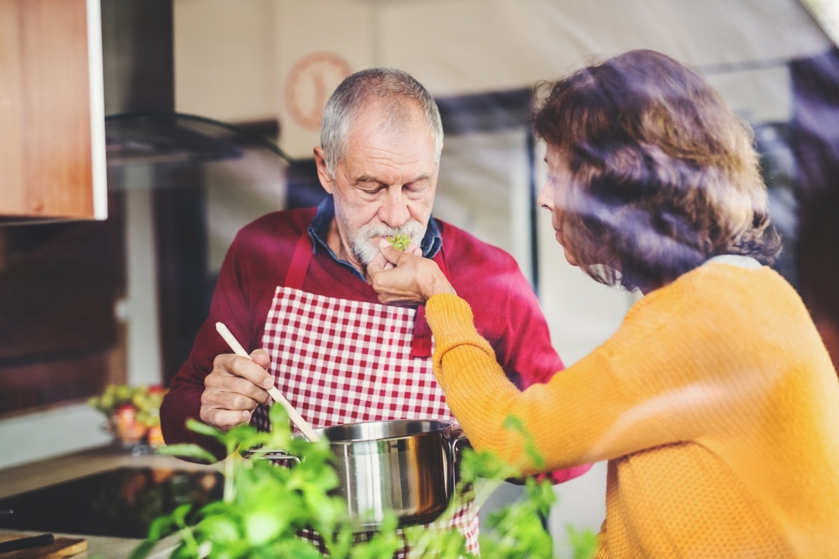 Senior couple preparing food in the kitchen. An old man and woman inside the house. Shot through glass.