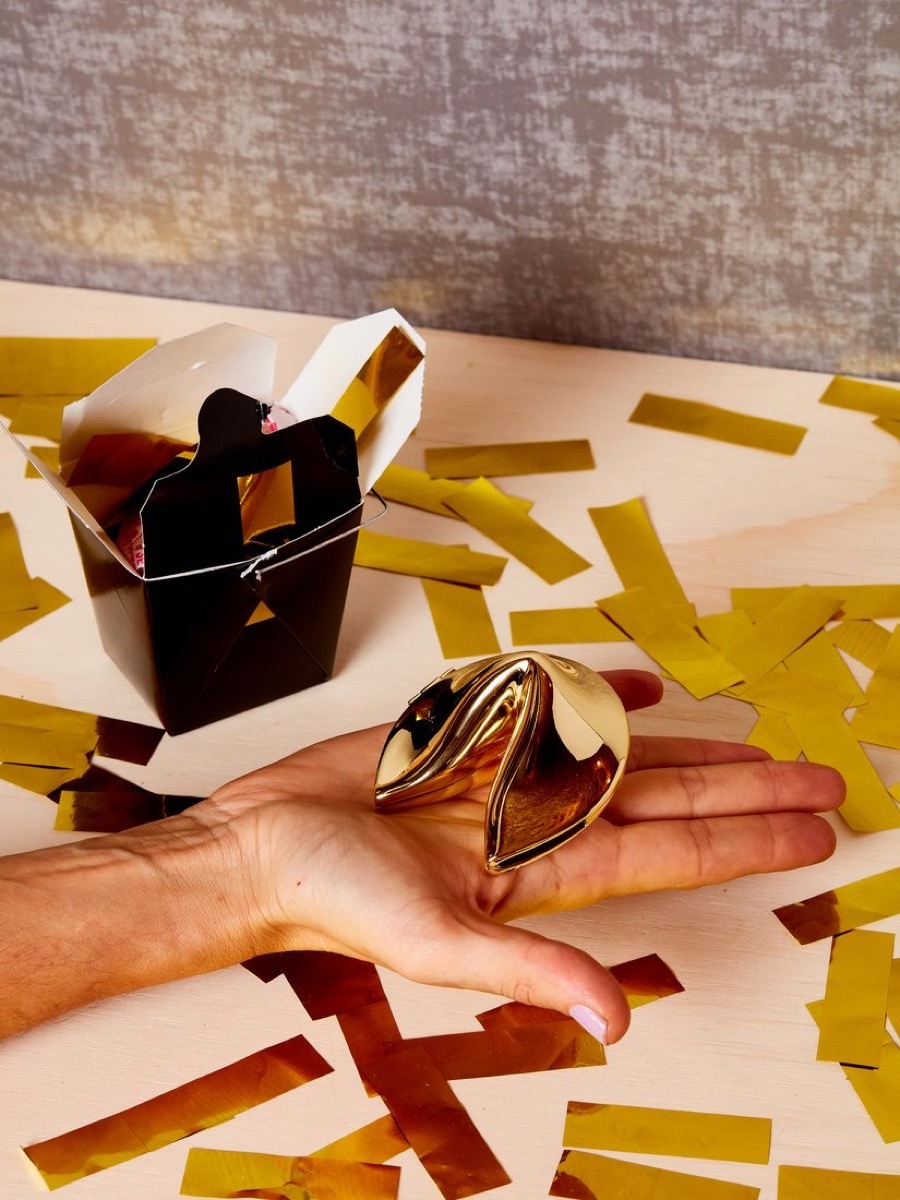 white hand holding gold fortune cookie with black takeout box and gold confetti in background