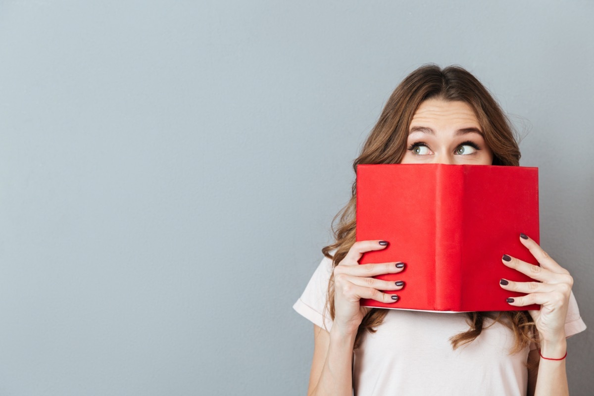 Portrait of a pretty young girl hiding behind an open book and looking away isolated over gray wall background