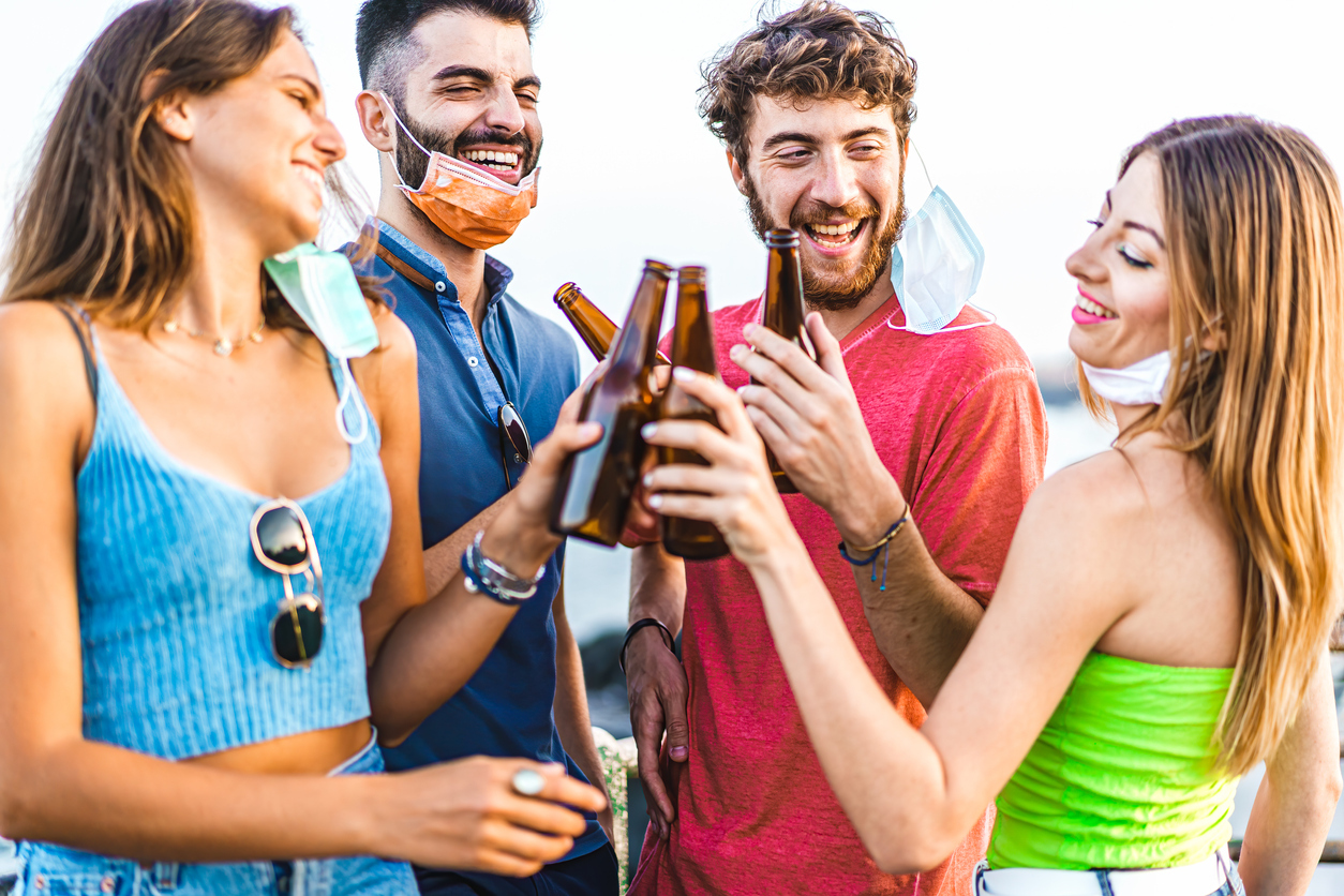 A group of four young men and women cheers beer bottles together with their face masks hanging off, making it easier to spread coronavirus