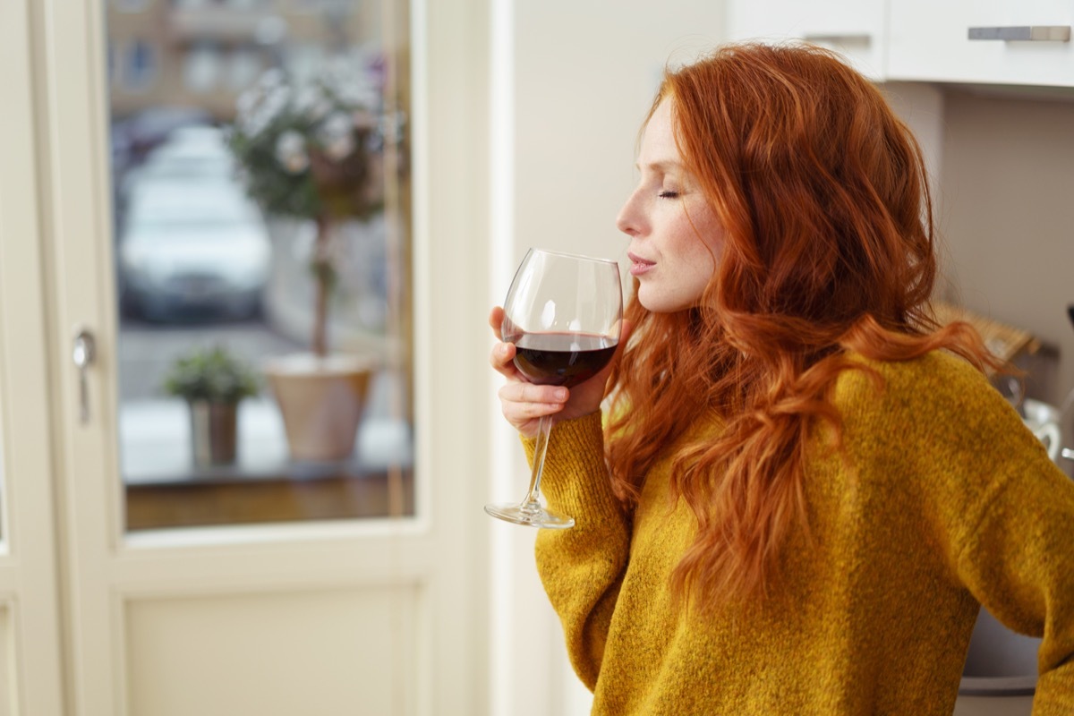 Young redhead woman standing in her apartment sipping a glass of red wine with her eyes closed in pleasure