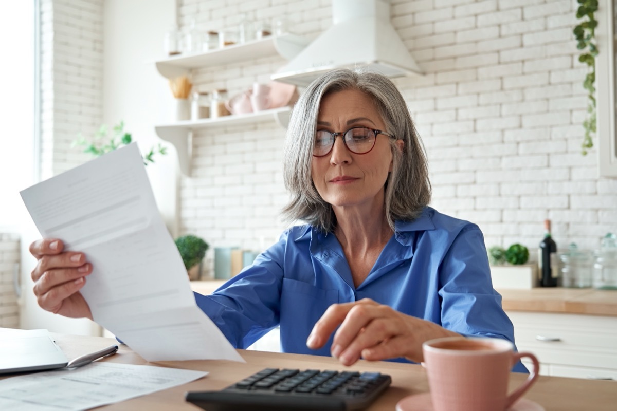 mature woman looking at a bill while using a calculator, sitting in her kitchen