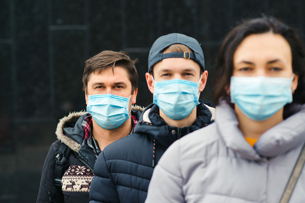 A group of three young people wearing face masks and winter coats stand in a line.