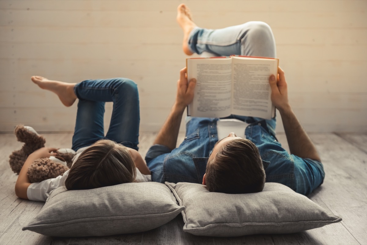 Charming little girl and her handsome young dad are reading a book while lying on the floor at home