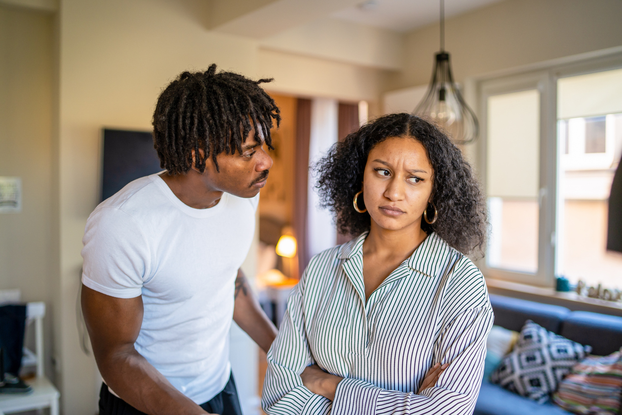 Woman is frowning and looking away from her boyfriend with her arms crossed, as he is looking worriedly into her face as they stand at home in their lounge