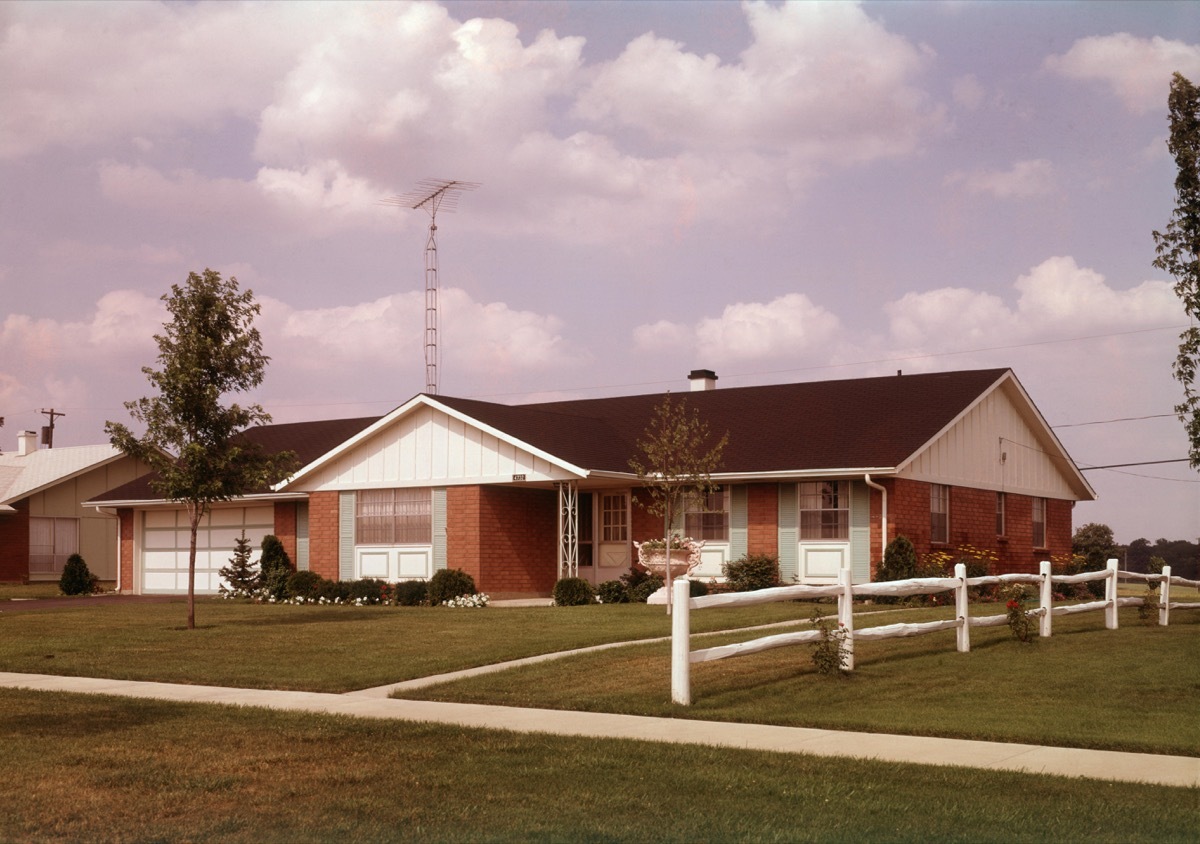 1960s huge suburban ranch home sits on green grass with white picket fence