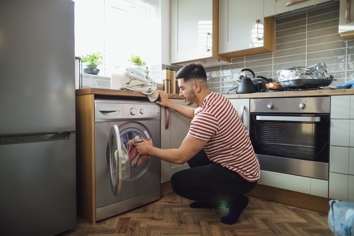 Man crouching down to put washing into his washing machine in his kitchen. He is in the Northeast of England.