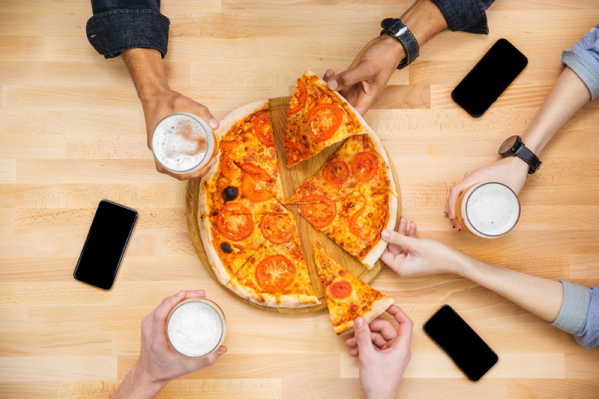 hands reaching for slices on pizza across table with phones and pints of beer on it