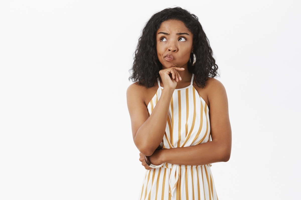 female in trendy striped yellow overalls smirking holding hand on chin in thoughtful pose gazing at upper left corner, thinking making choice