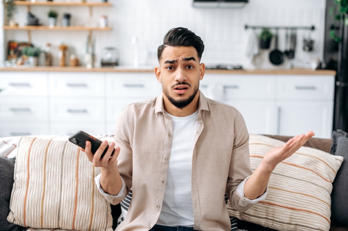 Man sitting on couch, holding phone and shrugging