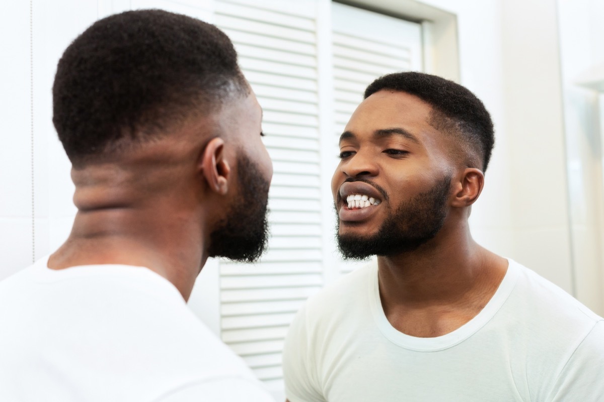 Young man looking in mirror at his mouth and teeth in bathroom, checking their health state