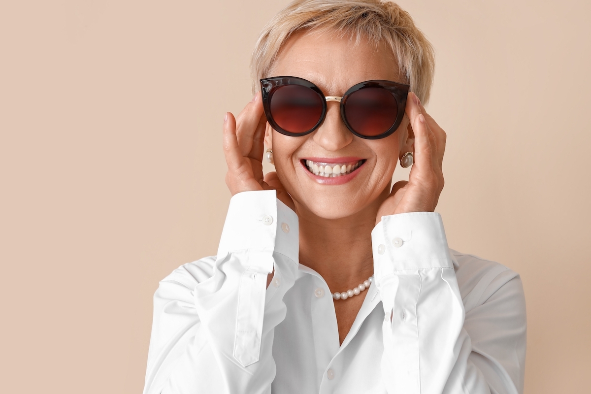 A stylish older woman wearing a white shirt, pearl jewelry, and large black sunglasses against a beige background