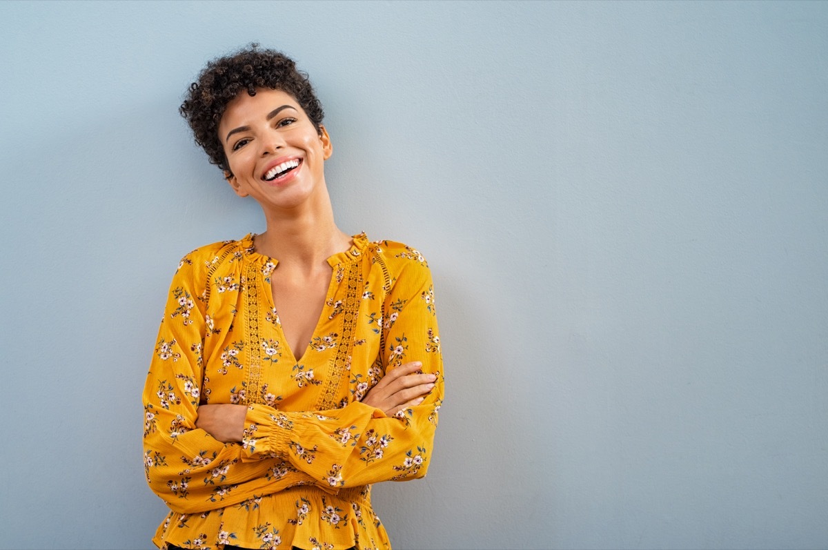 Portrait of beautiful cheerful girl smiling and looking at camera. Happy african woman in casual standing on blue background. Brazilian stylish woman with crossed arms and curly hair isolated with copy space.