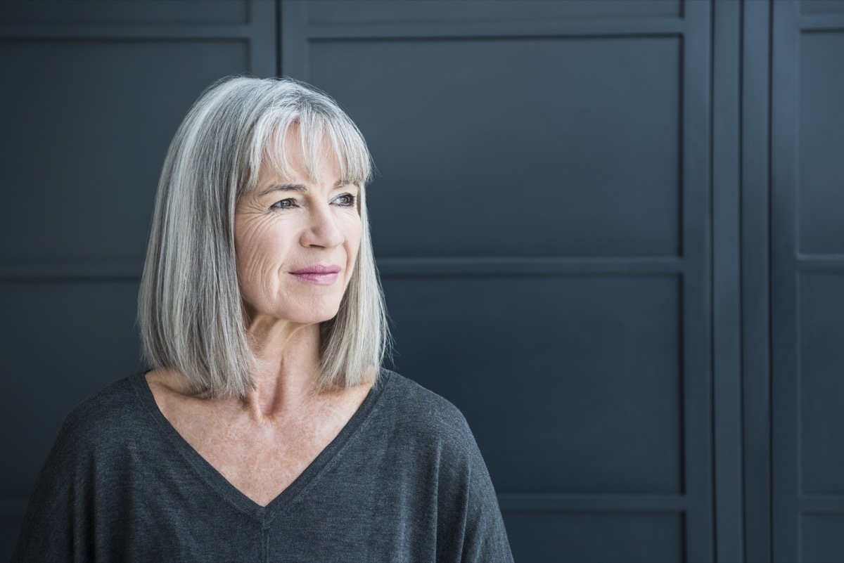 One senior woman with a grey bob in front of a dark grey background. Portrait of smart senior woman in her 60s, looking away from camera.