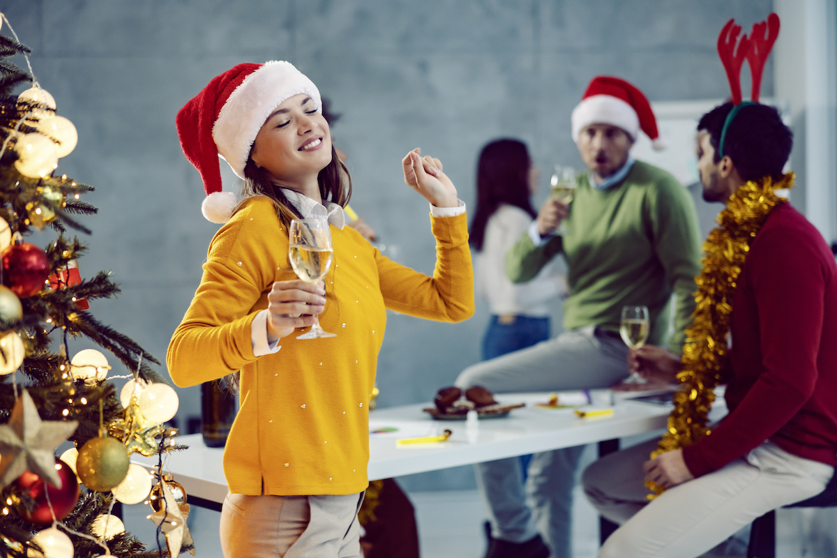 A young businesswoman is standing next to a Christmas tree in the boardroom with a glass of champagne in her hands and dancing alone.