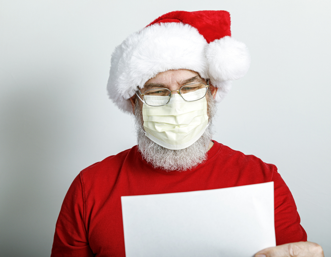 A Santa Claus character reads from a list while wearing a protective face mask