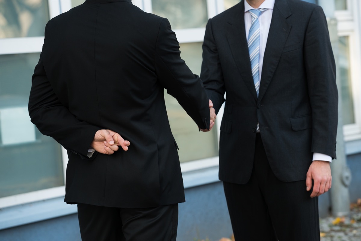 Midsection of dishonest businessman with fingers crossed shaking hands with partner outdoors