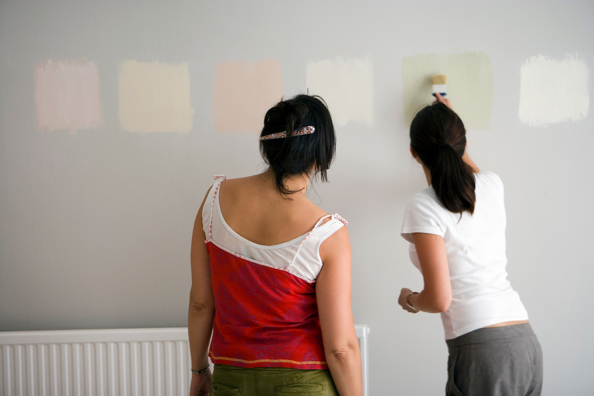 Rear view of two women testing different paint shades over the wall.