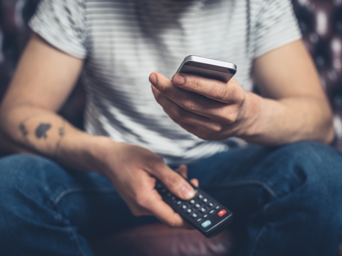 A young man is sitting on a sofa with a remote control and a smartphone