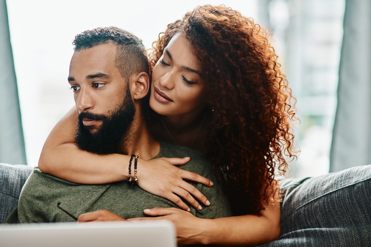 Shot of a young woman hugging her husband while he uses a laptop on the sofa at home