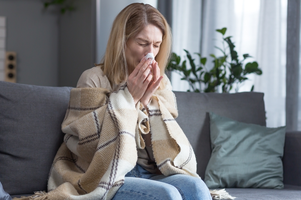 A woman blowing her nose while sick on the couch with COVID symptoms